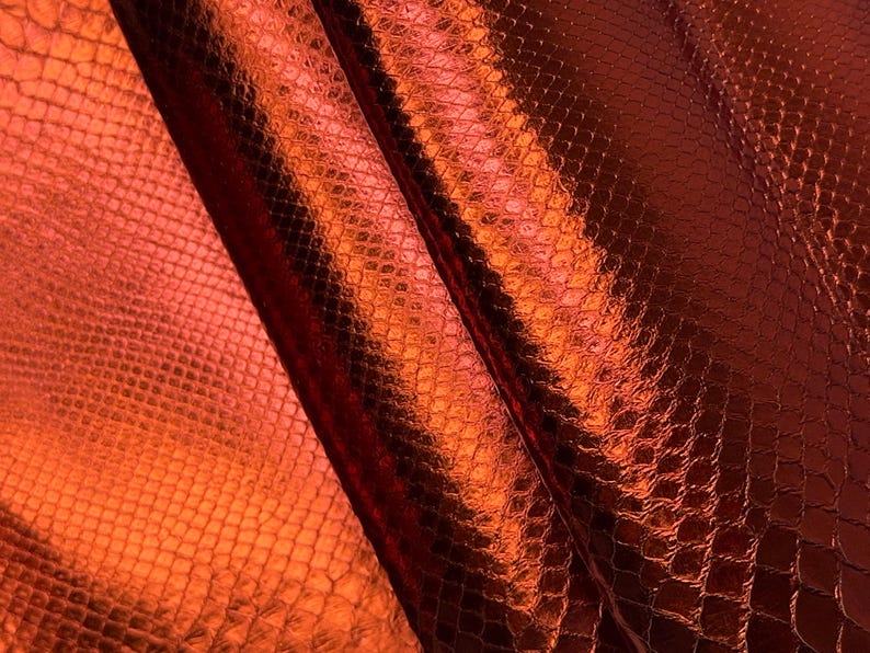 May include: Close-up of a textured, metallic orange-red fabric with a snakeskin pattern. The material has a glossy finish, reflecting light and creating highlights and shadows. The fabric is folded, showing the texture and depth of the material.