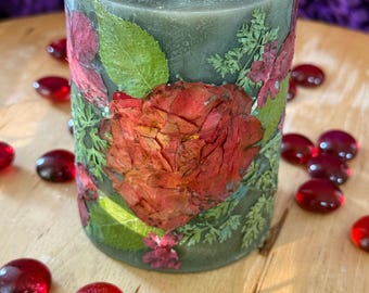 A large dark green unscented candle transformed with pressed flowers. A deep orange rose , azaleas and foliage adorn the candle.