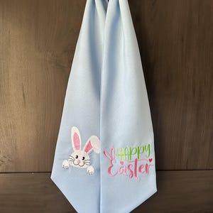 Puede incluir: Toalla colgante de Pascua de color azul claro. Presenta un diseño bordado de un conejito blanco y rosa y las palabras "Happy Easter" en verde y rosa. Cuelga de una puerta de madera.