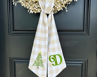 May include: A beige and white checkered door scarf with a Christmas tree and the letter "D" embroidered in green. The scarf is hanging on a black door with a white berry wreath.