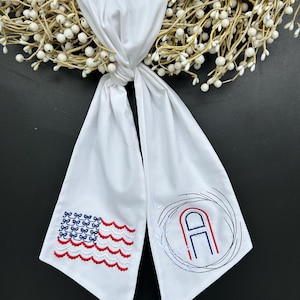 May include: White fabric sash with embroidered American flag design and the letter "A" inside a circular wreath. The sash is tied around a wreath of white berries and brown twigs, set against a black background.