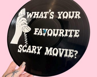May include: Black vinyl record with white text that reads "WHAT'S YOUR FAVOURITE SCARY MOVIE?" A white graphic of a hand holding a telephone is on the left side. The background is pink.