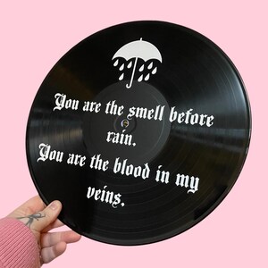May include: A black vinyl record with white text that reads "You are the smell before rain. You are the blood in my veins."  An umbrella with raindrops is above the text.