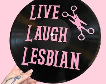 May include: Black vinyl record with the text "LIVE LAUGH LESBIAN" in pink lettering. A pair of pink scissors is also on the record.