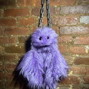 May include: A purple faux fur bag with a shaggy texture, featuring two small black bead eyes and a silver-toned metal chain strap. The bag has a rounded top section and a wider, flared base, creating a creature-like appearance with long fibers.