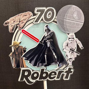 May include: A Star Wars themed cake topper featuring the number 70 in black glitter, the name Robert in black glitter, a Millennium Falcon, a Death Star, Yoda, a Stormtrooper, and Darth Vader holding a red lightsaber on a light blue circular background.