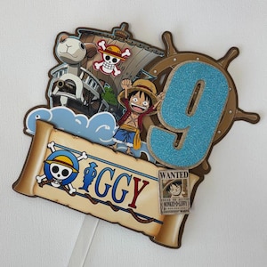 May include: A decorative cake topper featuring One Piece anime elements, including a ship, a character with a straw hat, a blue glittery number 9, and a scroll with the name Iggy and a small Wanted poster for Monkey D. Luffy.