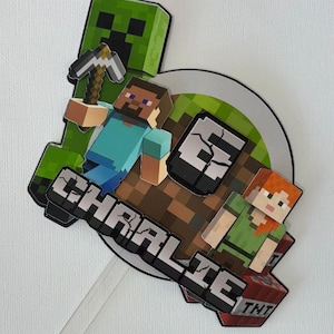 May include: A decorative cake topper featuring Minecraft characters including a green Creeper, Steve holding a pickaxe, and Alex. The design includes a brown dirt block, a TNT block, and the name CHRALIE in a blocky, pixelated white font.