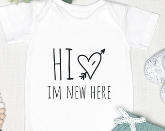 May include: White baby onesie with black text that reads "Hi I'm new here" with a heart and arrow graphic. The onesie is laid out on a white surface with a pair of white baby shoes, a green baby skirt, and a wooden bead teether.