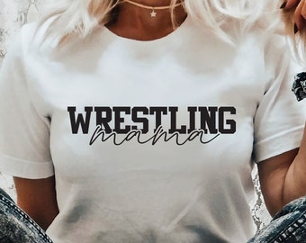 Może przedstawiać: Biała koszulka z napisem "WRESTLING mama" czarnymi literami. Koszulkę nosi osoba w jeansach z przetarciami.