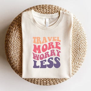 Peut inclure: Un t-shirt blanc avec le texte "TRAVEL MORE WORRY LESS" dans une police de caractères dégradée arc-en-ciel.