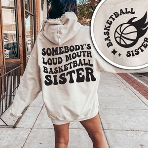 May include: A white hoodie with a black graphic that says "Somebody's Loud Mouth Basketball Sister" in a retro font. The graphic also includes a basketball with flames.