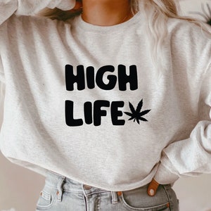 Peut inclure: Un sweat-shirt gris avec le texte "HIGH LIFE" en lettres majuscules noires et un motif de feuille de marijuana noire.