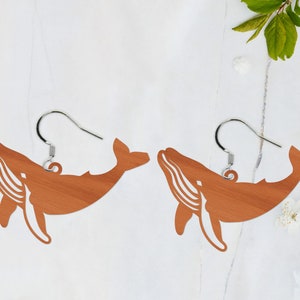 Może przedstawiać: Para drewnianych kolczyków w kształcie wieloryba z srebrnymi haczykami. Wieloryby są jasnobrązowe i mają szczegółowy zarys.
