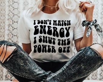 Könnte beinhalten: Weißes T-Shirt mit dem Text "I DON'T MATCH ENERGY I SHUT THE POWER OFF" in fetter, schwarzer Retro-Schrift. Die Person trägt eine Jeans aus Distressed-Denim.