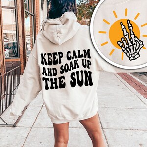 May include: A white hoodie with black text that reads "Keep Calm and Soak Up The Sun" in a retro font. The hoodie is being worn by a person standing on a sidewalk.