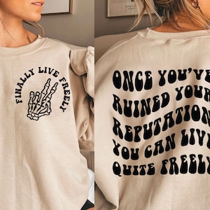 May include: A beige sweatshirt with a black skeleton hand graphic on the front and the text "Finally Live Freely" and the text "Once You've Ruined Your Reputation You Can Live Quite Freely" on the back.