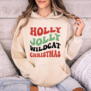 May include: A cream-colored hoodie with a red, green, and black graphic that says "HOLLY JOLLY WILDCAT CHRISTMAS".