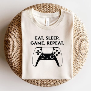 May include: Cream-colored t-shirt with the text "EAT. SLEEP. GAME. REPEAT." above a black and white controller graphic. The shirt is made of a soft material and is suitable for casual wear.