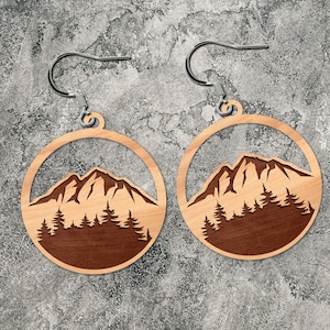 May include: A pair of wooden earrings with a mountain and pine tree design. The earrings are brown with a light wood grain and have silver hooks.