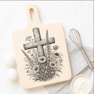 May include: A light-colored wooden cutting board with a handle featuring a black ink illustration of a rustic wooden cross surrounded by a dense arrangement of various wildflowers, including daisies and tall stalks, centered on the board surface.