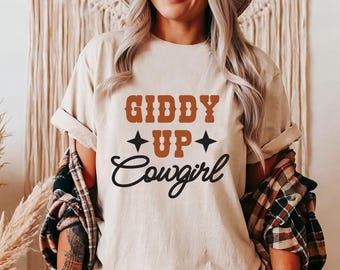 May include: A beige t-shirt with the text "Giddy Up Cowgirl" in brown and black lettering. The text is surrounded by two black stars.