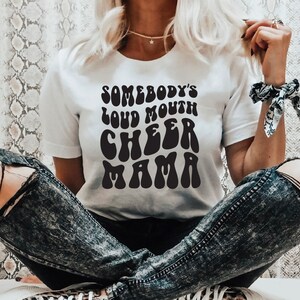 May include: White t-shirt with black text that reads "Somebody's Loud Mouth Cheer Mama" in a retro font.