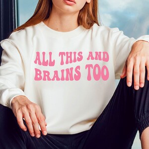 Puede incluir: Sudadera blanca con el texto "ALL THIS AND BRAINS TOO" en letras retro rosas.