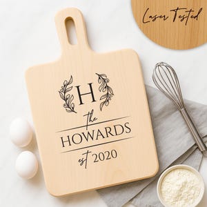 May include: A light-colored wooden cutting board with a handle, engraved with the letter "H" inside a floral wreath, the words "the HOWARDS est 2020". A whisk, eggs, and flour are next to the board. The text "Laser Tested" is on a wooden circle.