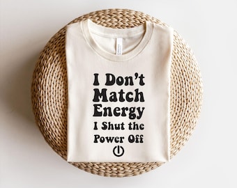 May include: Cream-colored t-shirt with the text "I Don't Match Energy I Shut the Power Off" in bold black letters. The design includes a power button symbol. The shirt is displayed on a woven, circular surface.
