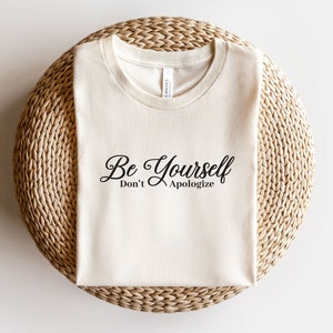 May include: A beige t-shirt with the text "Be Yourself Don't Apologize" printed in black.