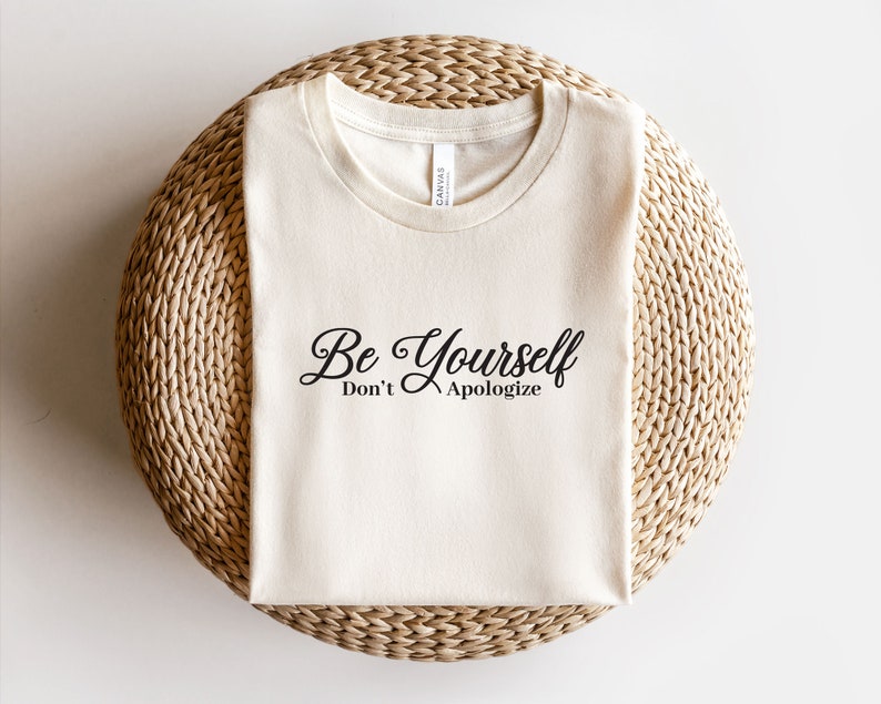 May include: A beige t-shirt with the text "Be Yourself Don't Apologize" printed in black.