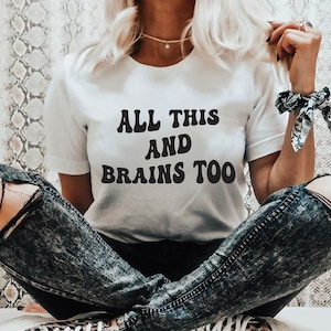 Puede incluir: Camiseta blanca con texto negro que dice "ALL THIS AND BRAINS TOO" en una fuente retro.