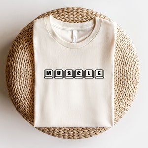 May include: A cream-colored t-shirt with the word "MUSCLE" spelled out in black letters, each letter appearing as a computer key.