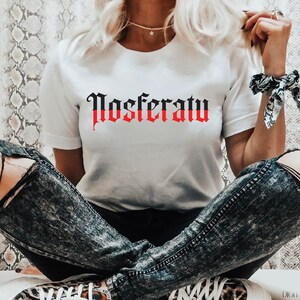 May include: White t-shirt with the word "Nosferatu" printed in black and red gothic lettering.