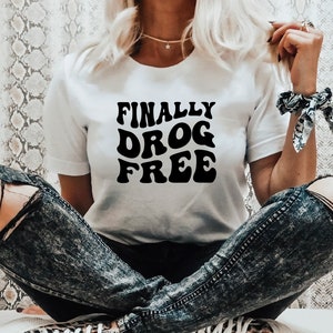 Puede incluir: Una camiseta blanca con texto negro que dice "FINALLY DRUG FREE" en una fuente retro.
