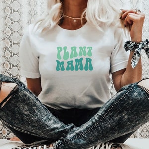 Peut inclure: Un t-shirt blanc avec le texte "PLANT MAMA" en police rétro verte et bleue.