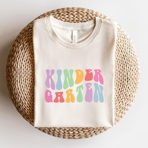 Può includere: Una maglietta color crema con la scritta "KINDERGARTEN" in lettere colorate in stile retrò. Il testo è nei toni del rosa, arancione, giallo, verde, blu e viola. La maglietta è appoggiata su una superficie circolare intrecciata.