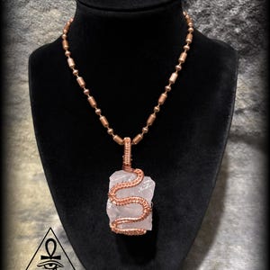 May include: A copper wire-wrapped necklace featuring a light pink, raw crystal pendant. The pendant is wrapped in a snake-like design. The necklace has a beaded chain and is displayed on a black bust.