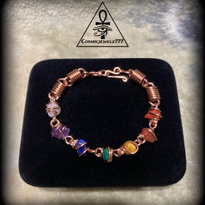 May include: A copper wire bracelet featuring seven colorful gemstones. The bracelet has a hook clasp and is displayed on a black velvet surface. The gemstones are wrapped in copper wire and are various colors.