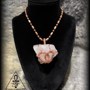 May include: A copper wire-wrapped quartz crystal pendant necklace. The raw, light pink crystal is encased in intricate copper wire, suspended from a beaded copper chain. The necklace is displayed on a black bust against a textured background. The text "CosmicJewelz777" is visible.