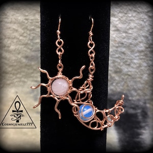 May include: A pair of wire-wrapped earrings featuring a sun and moon design crafted from copper-colored metal. The sun charm holds a pale pink round stone, while the crescent moon charm contains a translucent blue stone. Includes a logo with an ankh.