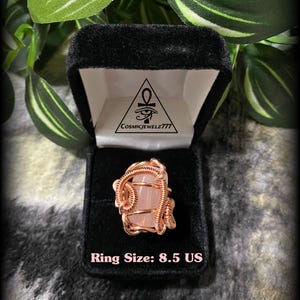May include: A rose quartz ring, size 8.5 US, displayed in a black velvet ring box. The ring features a light pink stone wrapped in copper wire. The box is set against a backdrop of green leaves.