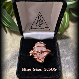 May include: A rose quartz ring with copper wire wrapping, displayed in a black velvet ring box. The ring is size 5.5 US. The box has a logo with an eye symbol and the text "CosmicJewelz777".