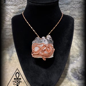 May include: A clear quartz crystal pendant necklace with copper wire wrapping. The pendant is displayed on a black mannequin. The necklace features a copper chain. The pendant is in the shape of a crystal cluster.