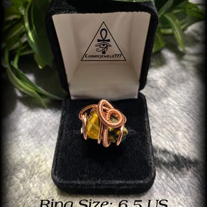 May include: A handcrafted ring featuring a tiger's eye stone wrapped in copper wire, presented in a black velvet box. The ring size is 6.5 US. The box has a logo with an eye symbol and the text "CosmicJewelz777".