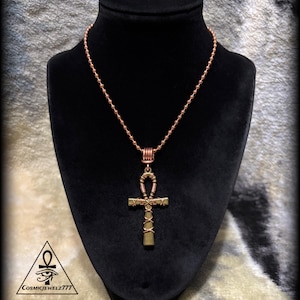 May include: A copper-colored ball chain necklace featuring an ankh pendant wrapped in thin copper wire. The pendant has a metallic bronze finish. A small logo with an ankh symbol and the text COSMICJEWELZ777 is visible in the bottom corner.