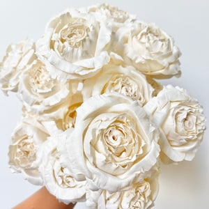 Könnte beinhalten: Ein Strauß weißer Sola-Holzrosen. Die Rosen sind in einer Hand angeordnet, wobei die Stiele nach unten zeigen. Die Rosen sind aus Sola-Holz gefertigt, einem natürlichen Material, das leicht und strapazierfähig ist.