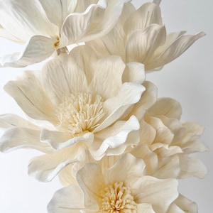 May include: Close-up of several large, cream-colored paper flowers. The flowers have multiple layers of delicate petals and textured centers. The arrangement is set against a plain white background, highlighting the intricate details of the floral design.