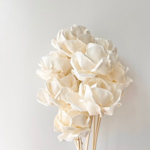 May include: A bunch of ivory-coloured artificial flowers, resembling roses, against a white backdrop. The flowers have multiple layers of petals and are attached to thin, light-brown stems. The arrangement is simple and elegant.